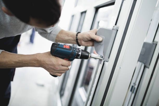 Person using a cordless drill to install a hinge on a window frame in a workshop setting.