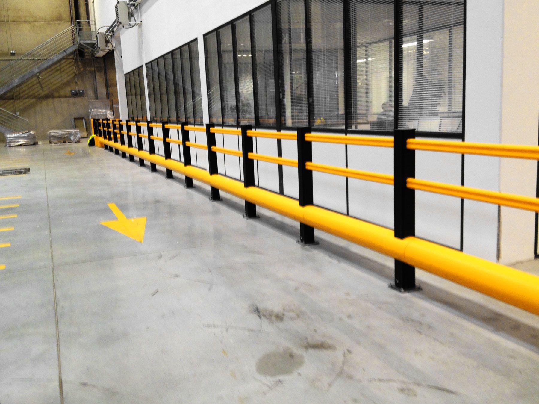 Interior view of a warehouse with yellow safety barriers and arrows on the floor, beside large windows.