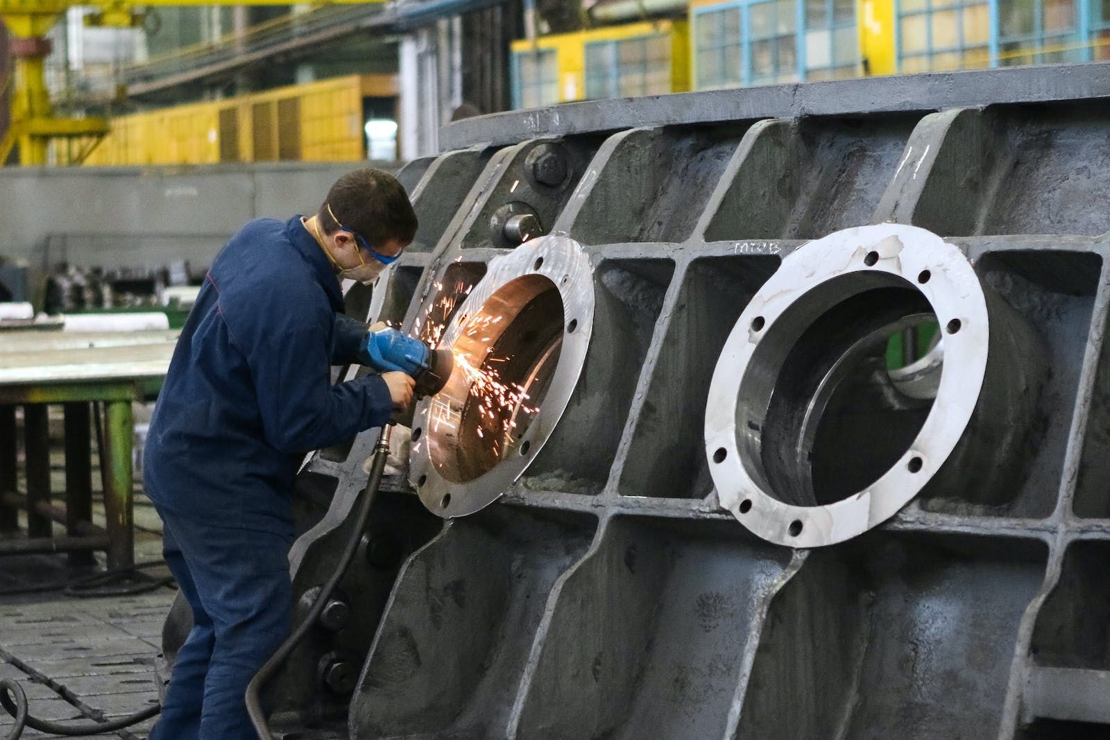 Worker in safety gear grinding a large metal structure with sparks flying in an industrial workshop.